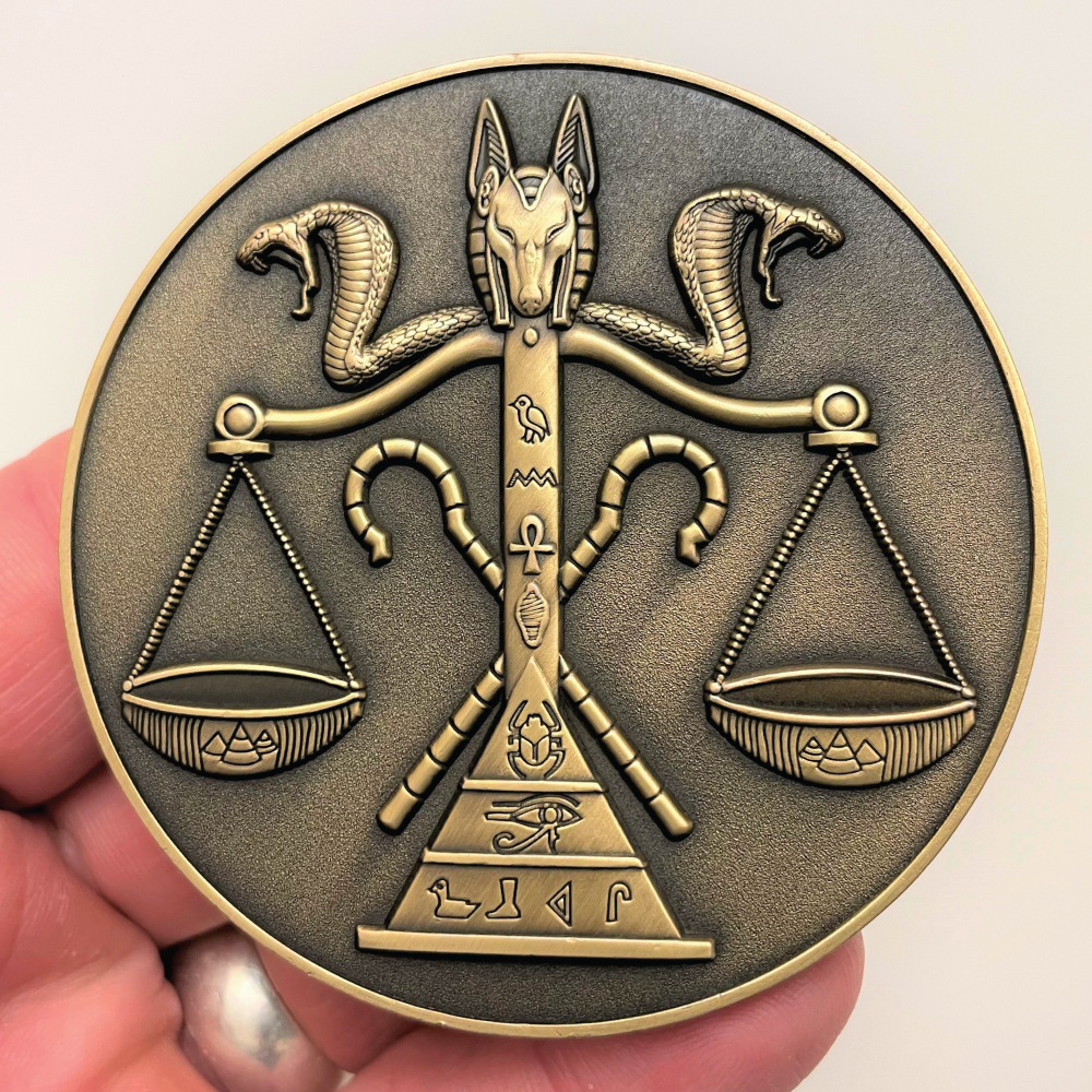 Gold metal coin in hand showing jackal head and cobras on top of scale with Egyptian symbols on it